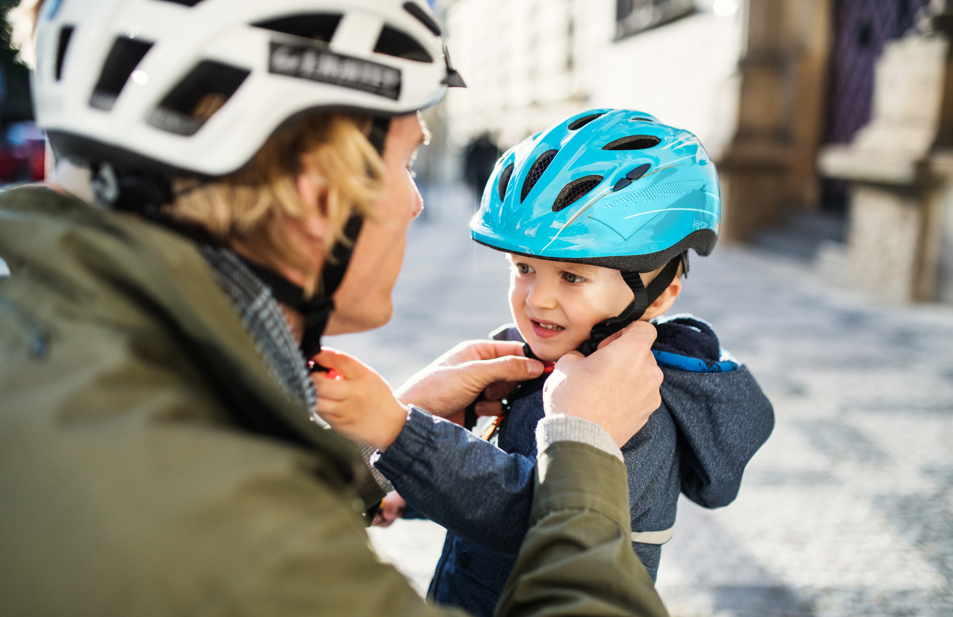 A young father putting on a helmet on his toddler son’s head outdoors in city.